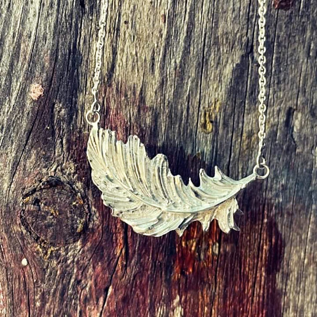 A silver necklace in the shape of a leaf in front of the bark of a tree.